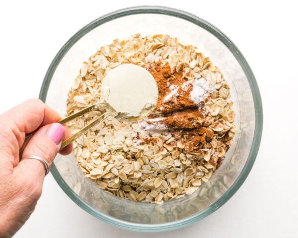Protein powder is being added to a bowl with oats, cinnamon, and more dry ingredients.