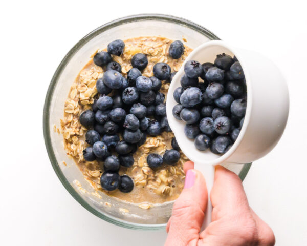 A cup of blueberries is being poured into a bowl with an oatmeal batter.