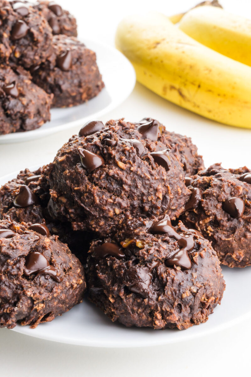 A plate of chocolate banana cookies sits in front of a stack of bananas.