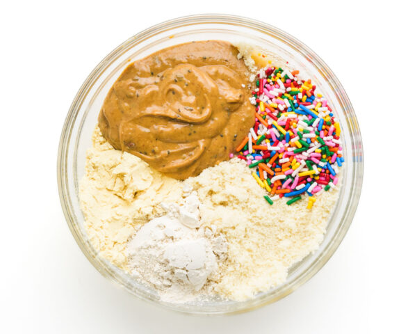 A glass bowl containing various ingredients: brown nut butter, flour, and colorful sprinkles.
