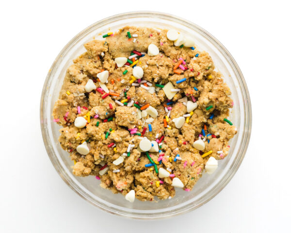 Bowl of cookie dough with colorful sprinkles and white chocolate chips on a white background.
