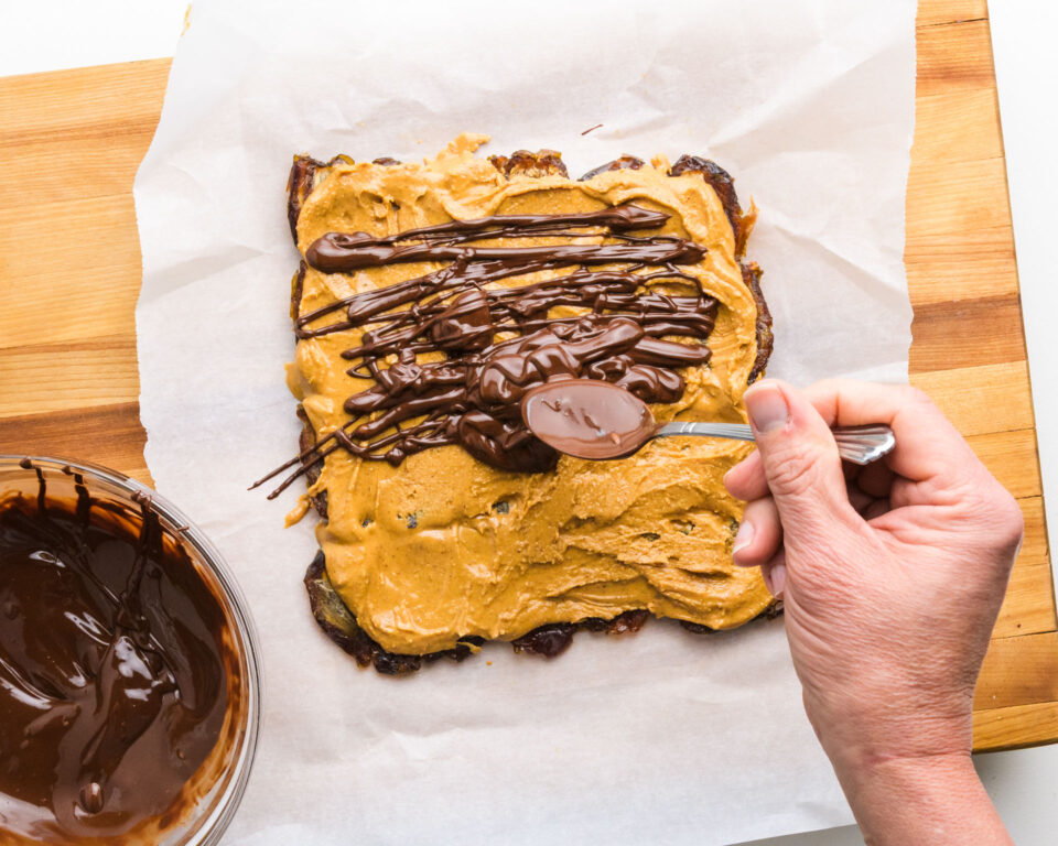 Melted chocolate is being drizzled over peanut butter on top of a layer of pressed medjool dates.