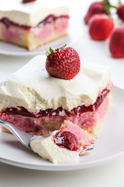 Strawberry shortcake cake slice topped with fresh strawberry and whipped cream, served with a fork on a white plate.