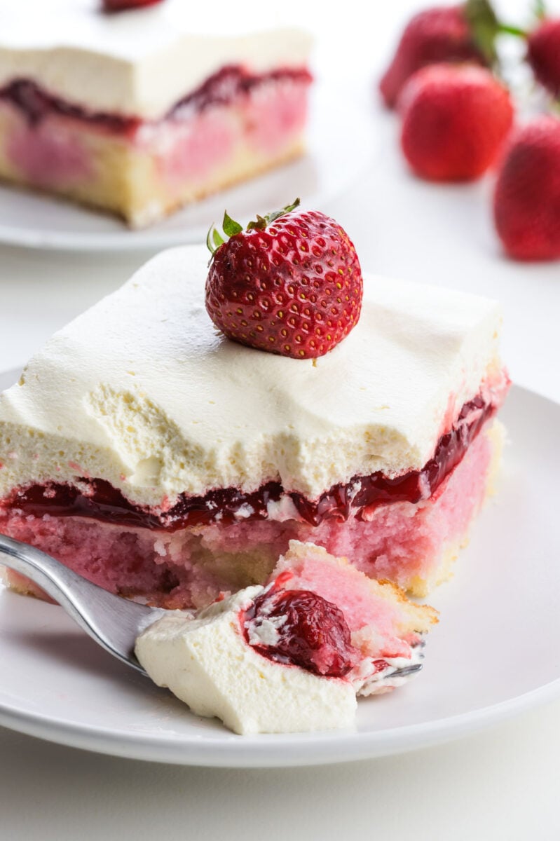 Strawberry shortcake cake slice topped with fresh strawberry and whipped cream, served with a fork on a white plate.