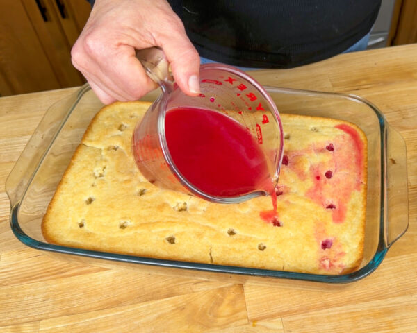 A hand pours bright red vegan jello over a baked cake with holes in a glass dish.