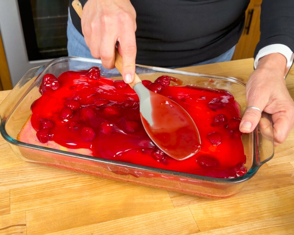 A hand using a spatula to spread red strawberry pie filling over a layer of cake in a glass dish on a wooden countertop.