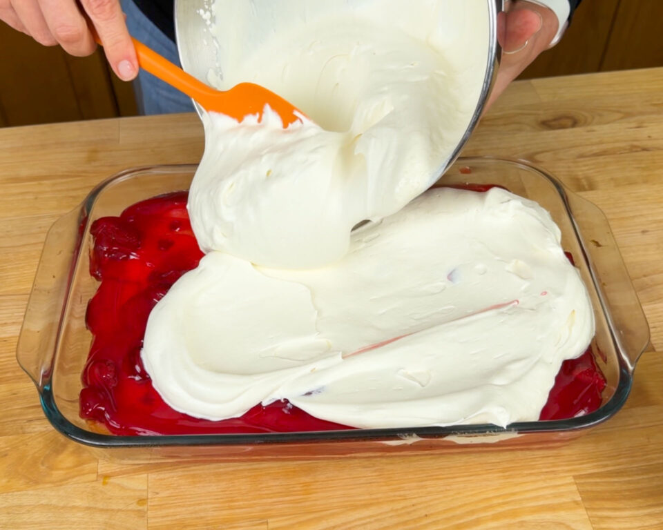Creamy topping being poured over a strawberry-covered cake in a glass dish on a wooden table.
