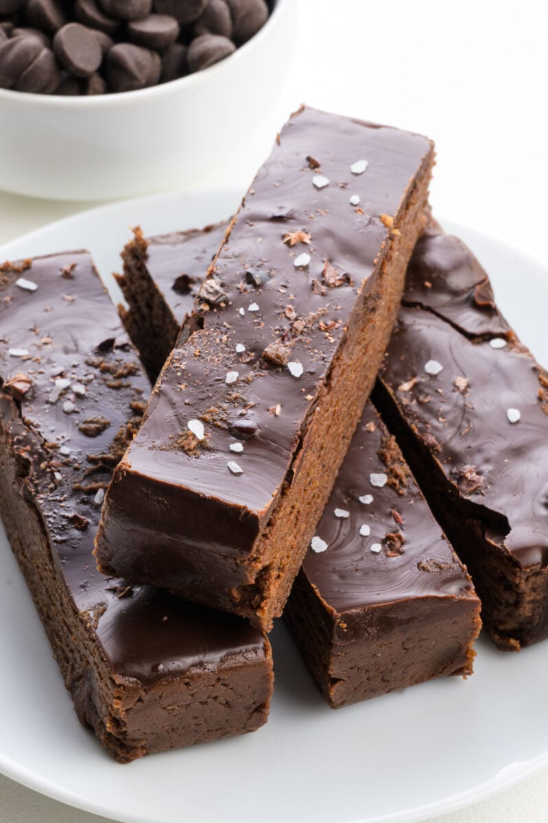Chocolate protein bars topped with sea salt, served on a white plate with chocolate chips in a bowl in the background.