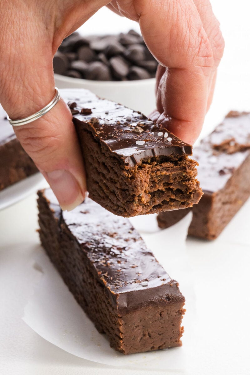 A hand holding a Chocolate Brownie Protein Bar with a shiny topping, surrounded by other chocolate bars and chocolate chips.