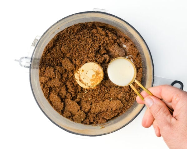 Hand holding a gold measuring spoon above a food processor filled with chocolate protein powder mixture.