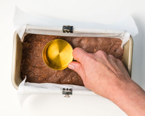 A hand holds a gold measuring cup over a protein bar slab in a parchment-lined pan.