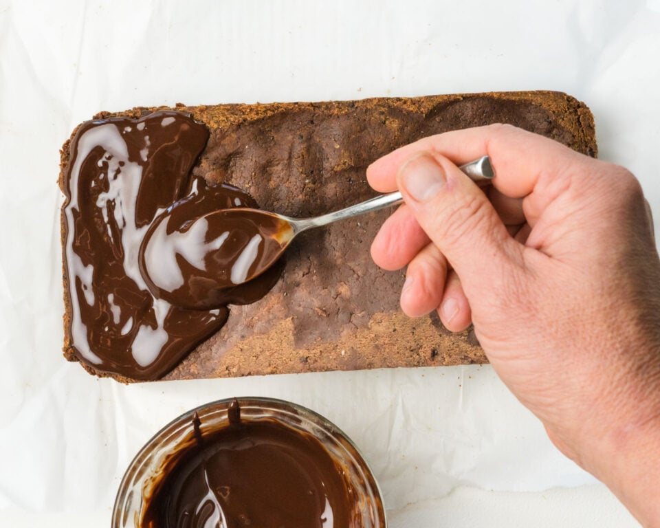 Hand spreading chocolate icing on a protein bar slab, with a jar of chocolate icing visible nearby.