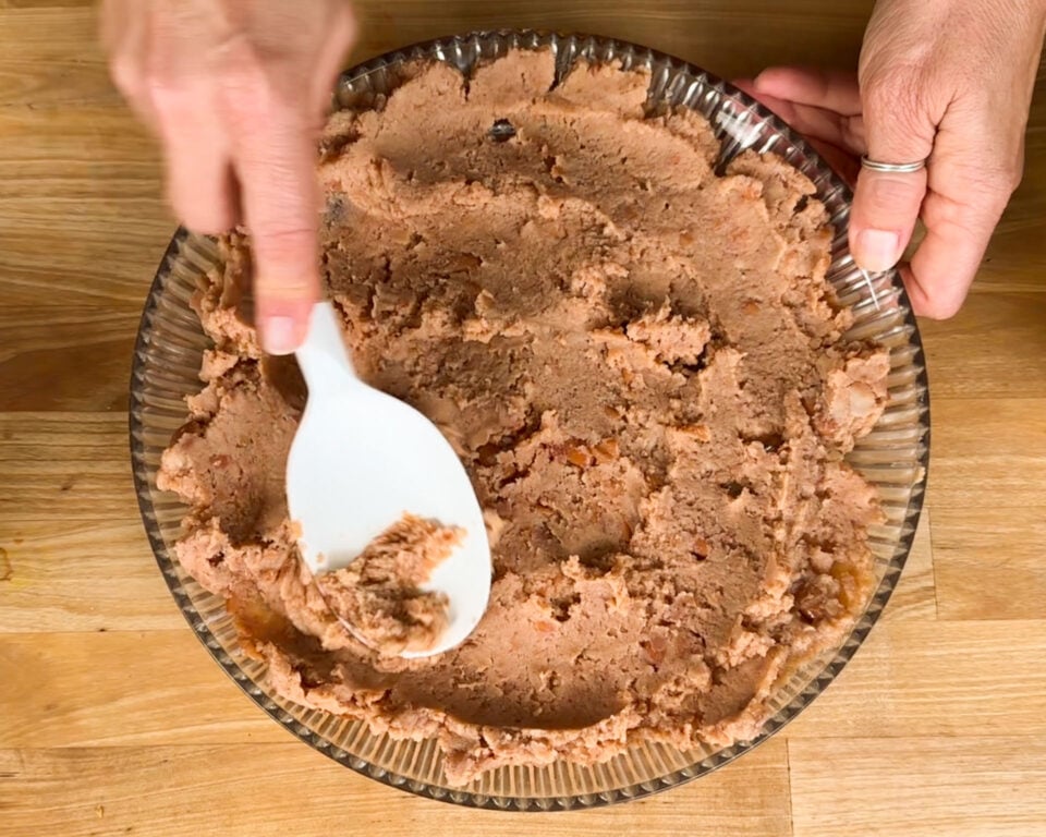 Hands spreading a mixture into a glass pie dish using a white spatula on a wooden surface.