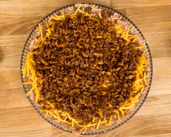A glass pie dish filled with layers of ground meat, shredded cheddar cheese, and a crispy topping.