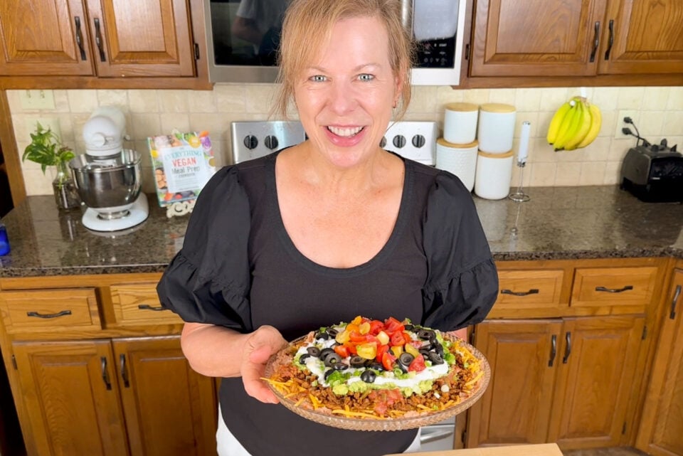 Marly in a black shirt holding a colorful vegan dish with toppings in a kitchen setting.