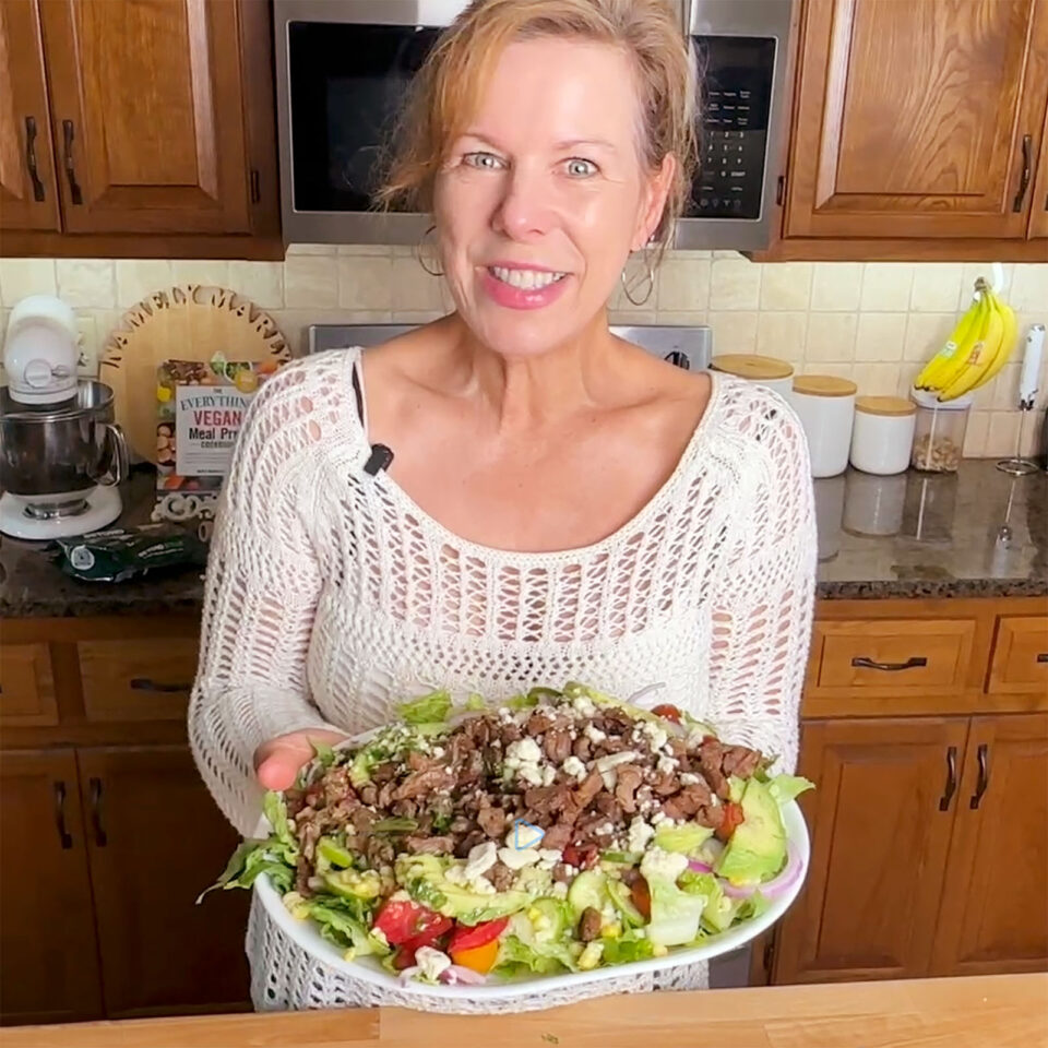Marly smiles while holding a large salad with mixed greens, toppings, in a kitchen with wooden cabinets.