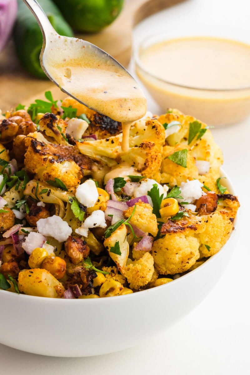 A bowl of roasted cauliflower salad topped with herbs, onions, corn, and a creamy dressing being drizzled from a spoon.