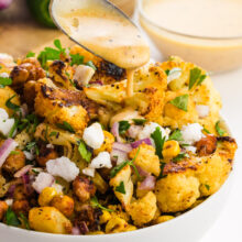 Roasted cauliflower salad with chickpeas, red onion, herbs, and a creamy dressing being drizzled over it.