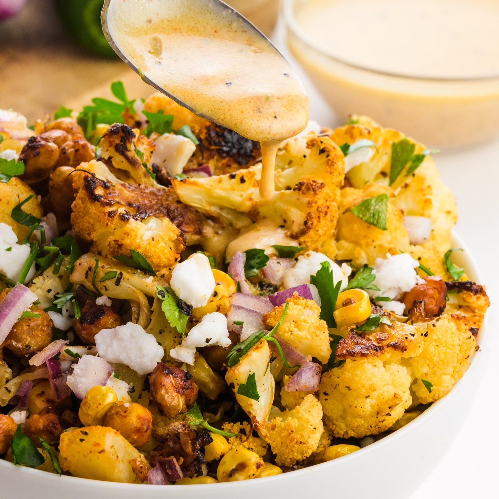 Roasted cauliflower salad with chickpeas, red onion, herbs, and a creamy dressing being drizzled over it.