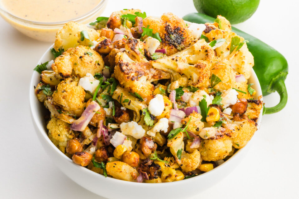 Bowl of roasted cauliflower mixed with chickpeas, onions, corn, parsley, and lime, with a sauce on the side.