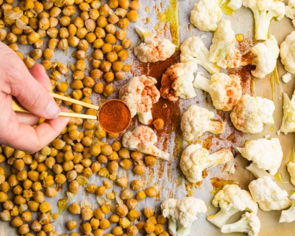 Hand using measuring spoons to sprinkle paprika over roasted cauliflower and chickpeas on a baking sheet.